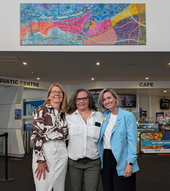 Three women stand in front of colourful painting