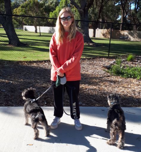 Woman with red jumper stands at dog park with two small dogs beside her