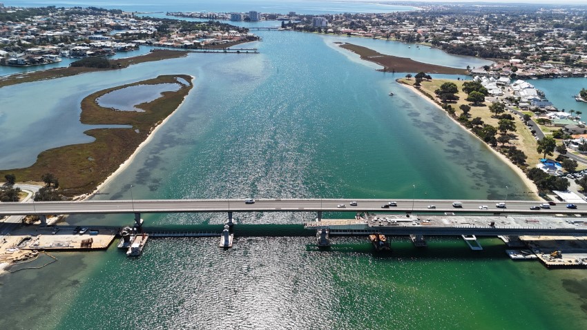 Daytime aerial image showing the Mandurah Estuary Bridge Duplication with the new bridge construction halfway across the Mandurah Estuary. The image shows the vast waterways and the coastal land on the sides.