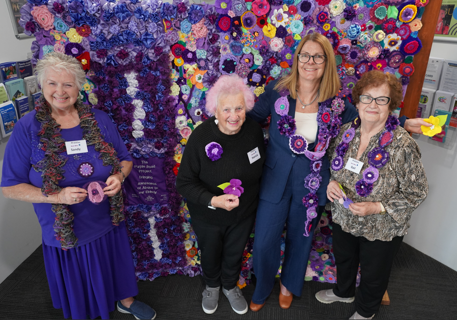 Knitting group members stand side by side with Mayor, in front of purple knitted artwork