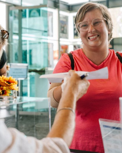 A smiling person hands a piece of paper to a staff member at an information desk inside a bright, modern building