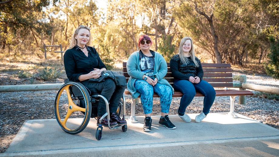 Three people sitting side-by-side, one lady on a wheelchair and two on a bench in a bushland setting.