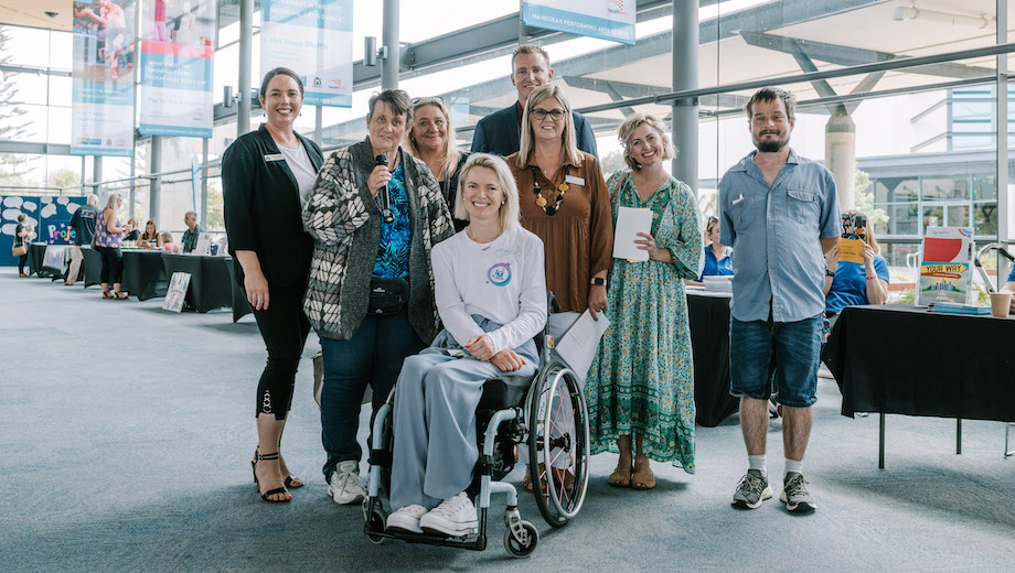 A group of eight people smiling. There are a range of ages, gender and disability represented in the group. One person is holding a microphone.