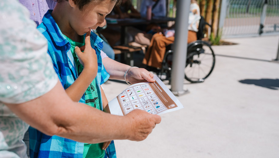 A young person is looking at a communication board which has pictures and text. There is an adult on each side of the young person and there is a picnic table in the background.