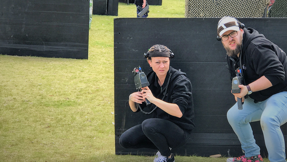 Two people hiding from others behind a black wall, playing laser tag.