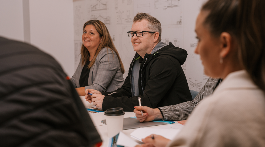 People sitting at a table during a professional workshop or meeting. A man and woman in the centre are smiling and listening. Other participants are seated with notebooks and coffee cups in front of them. 