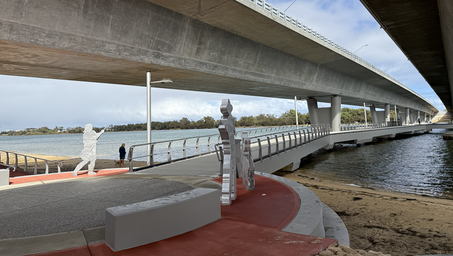 Accessible fishing platform under a large bridge with a wide ramp, railings and public artwork beside the water.