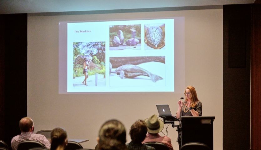 A woman stands behind a lectern, speaking to a group of people seated in a lecture hall. behind her is a projected screen showing different pieces of public art.