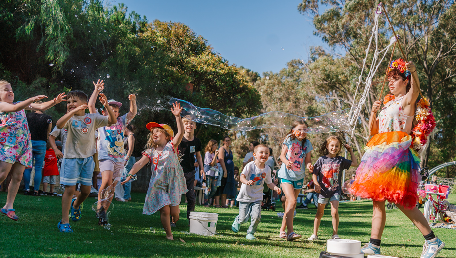 A group of children in the park running and playing with bubbles that a young lady dressed as a fairy is releasing.