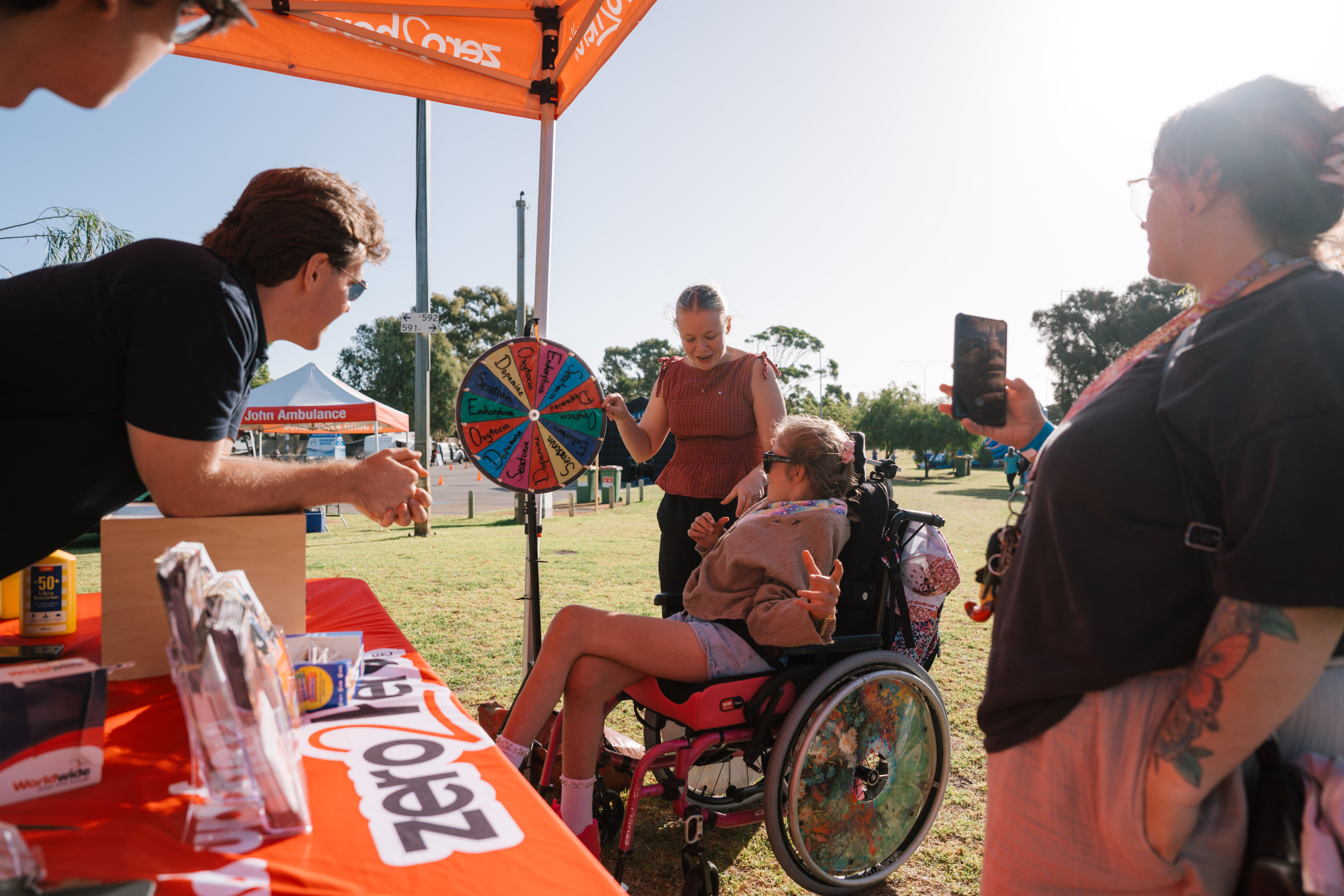 A person who uses a wheelchair takes part in a prize wheel activity at an outdoor community event, surrounded by several people under an orange marquee.