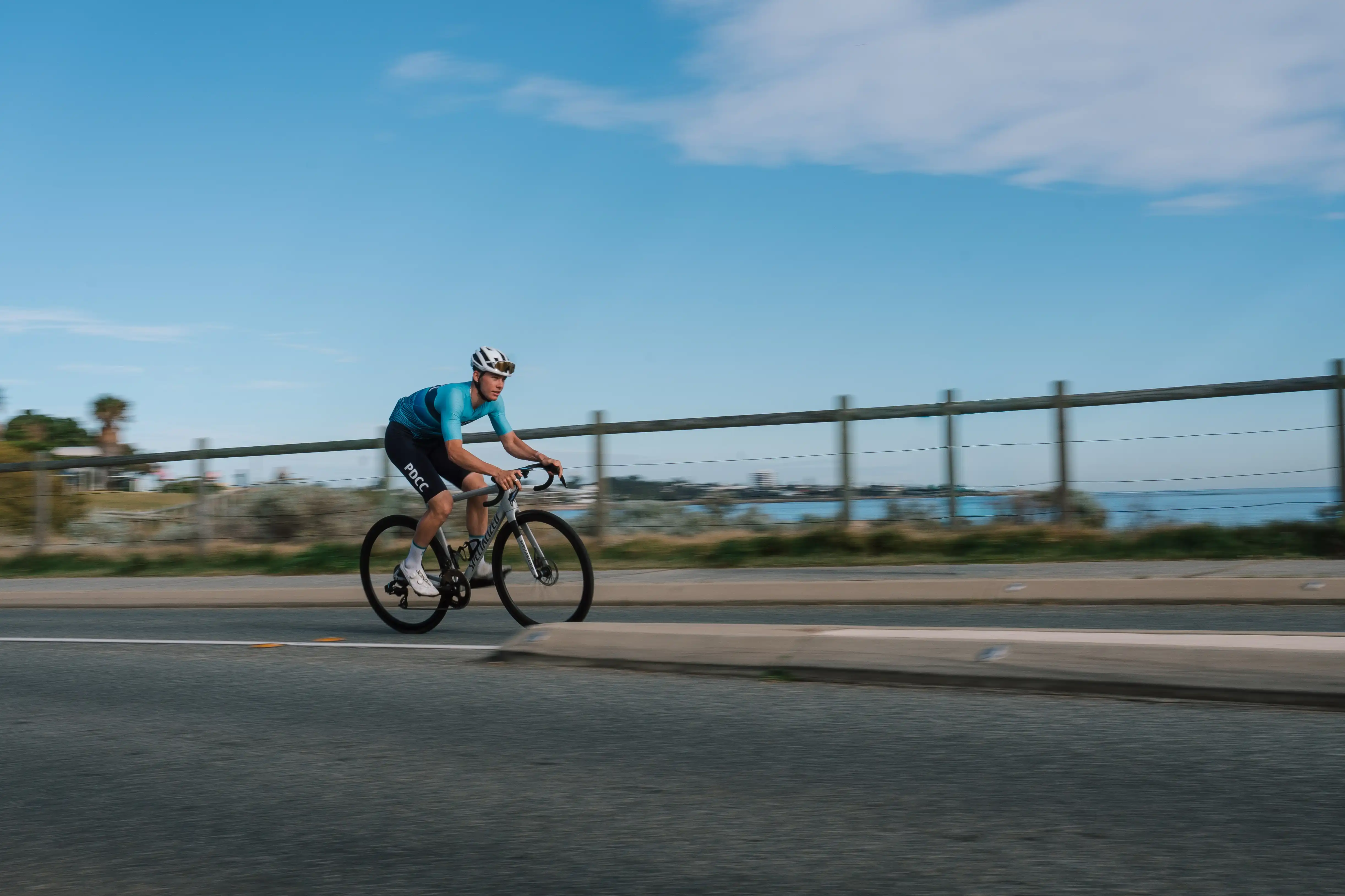 Cycling athlete riding a road bike along a coastal road, with ocean visible in the background.