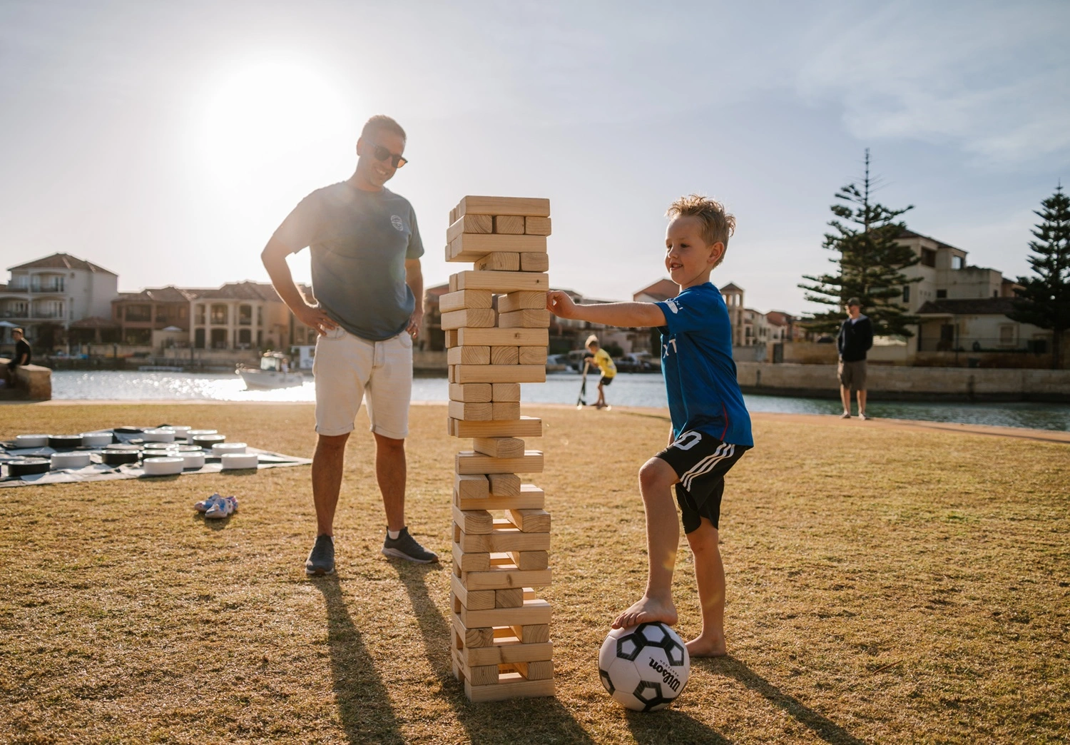A young boy with a blue tshirt and black shorts with one foot on top of a soccer ball, playing giant jenga with a family member at a Music in the Burbs event.