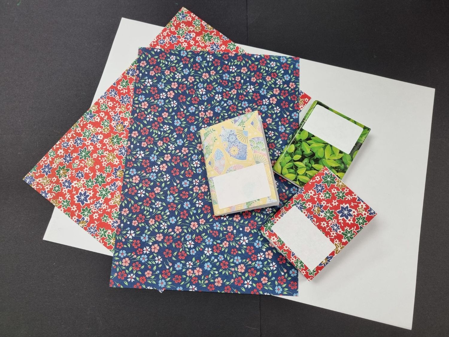Three handmade miniature notebooks with blank white labels sit on top of sheets of vibrant, floral-patterned craft paper laid over a plain white background