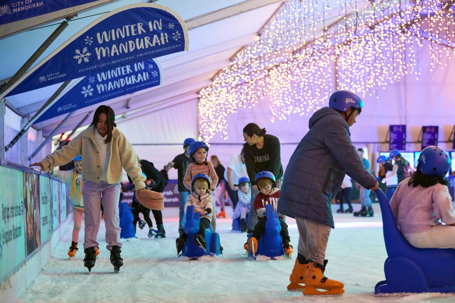 A busy indoor ice skating rink decorated with bright fairy lights and banners. Adults and young children in helmets are skating, with some toddlers using blue plastic seal-shaped skating aids.