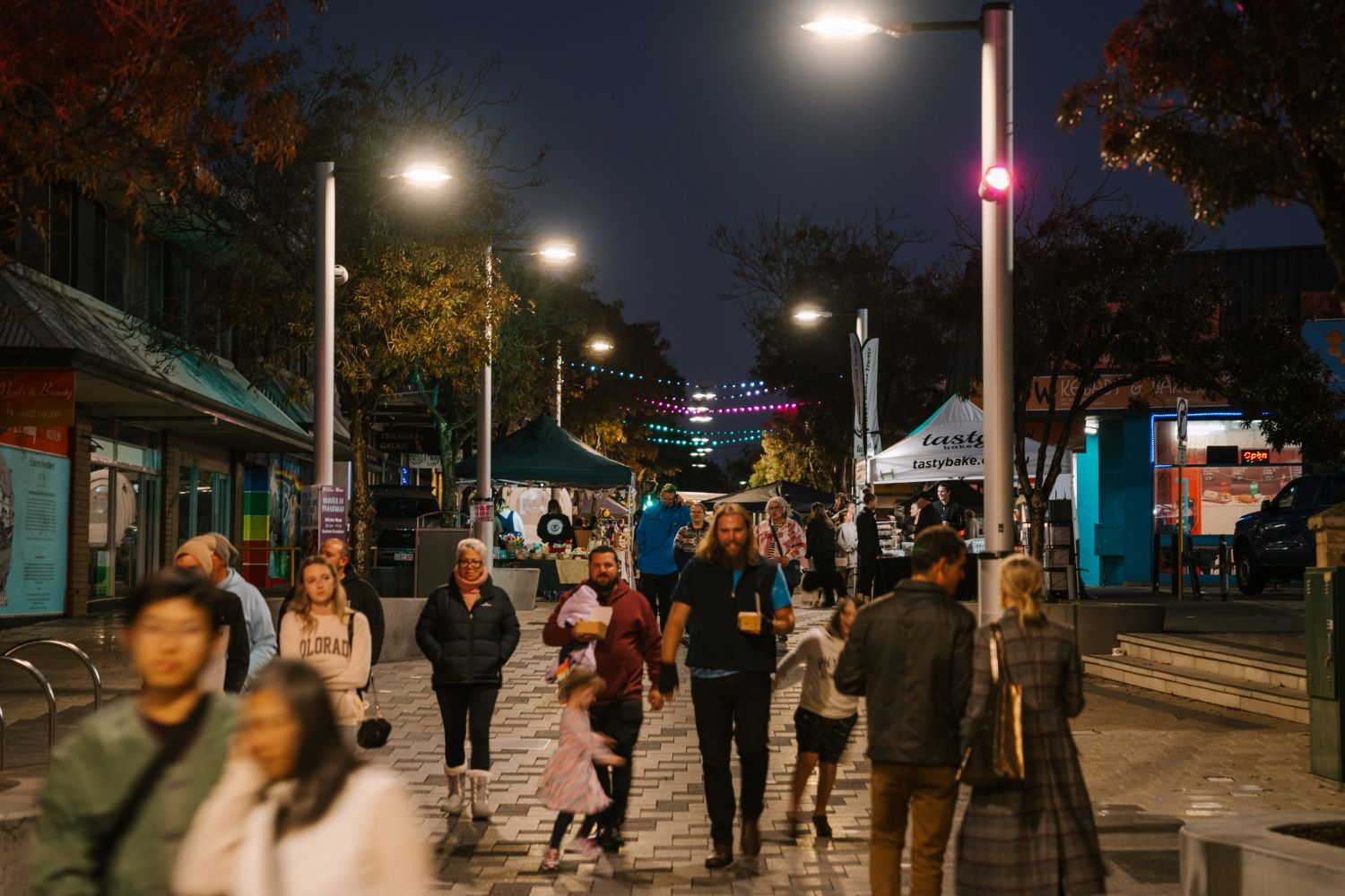 A vibrant night market on a paved pedestrian street illuminated by overhead streetlights and colorful string lights. Diverse crowds of people stroll past outdoor market stalls, including a food tent.