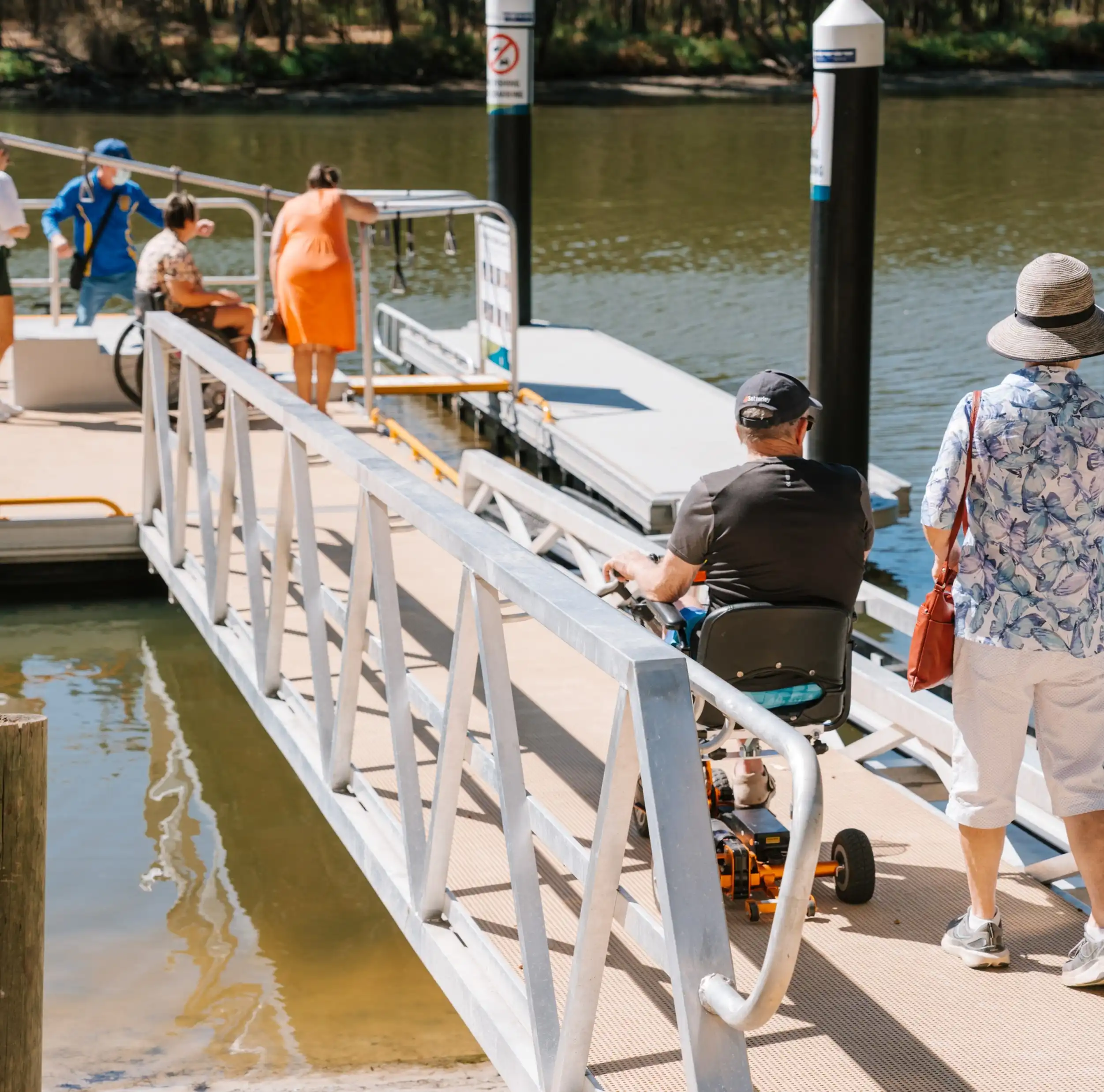Person using a mobility scooter travelling down an accessible jetty ramp toward an accessible paddle launch facility while others wait nearby.