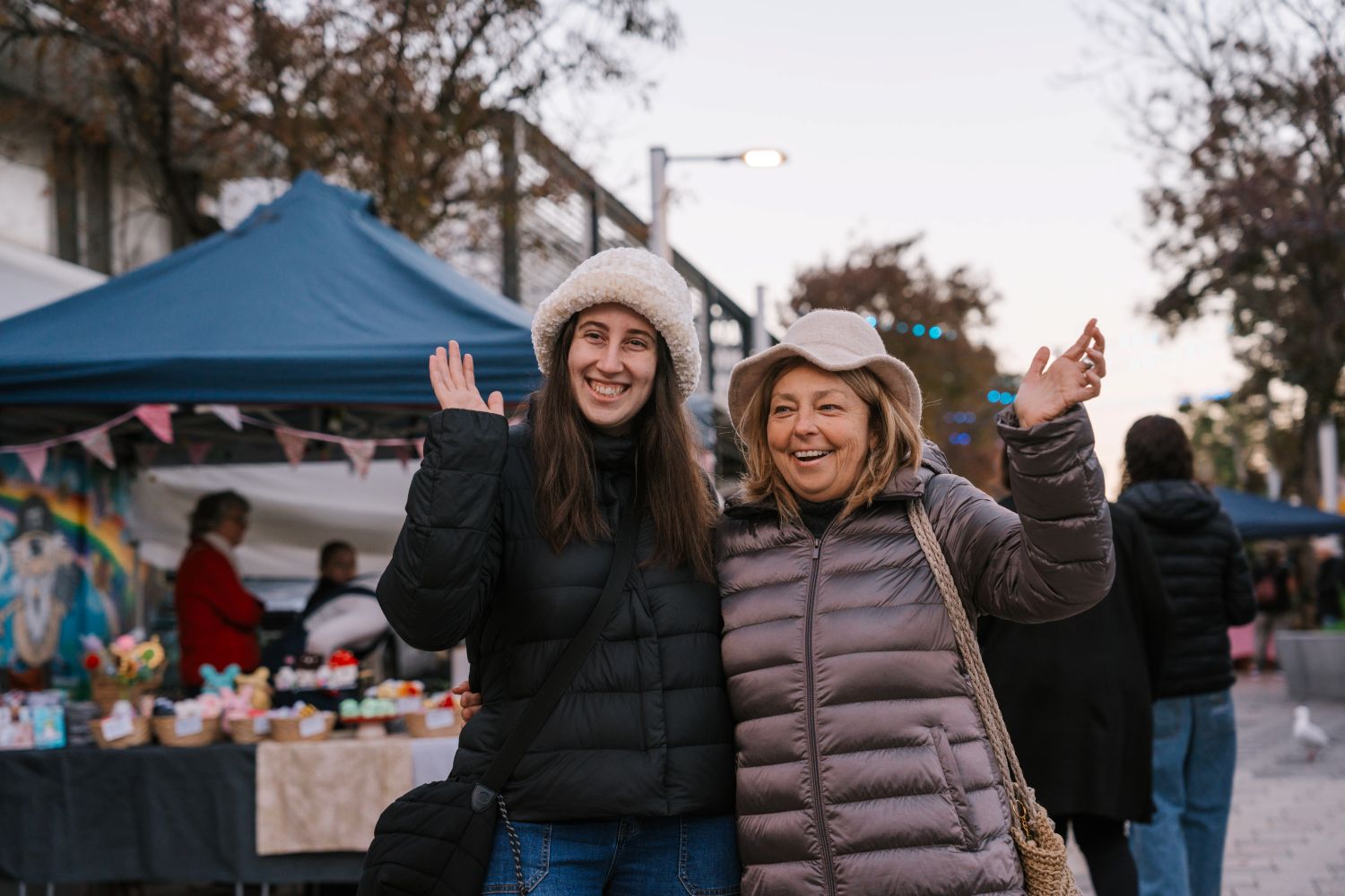 Two smiling women in winter coats and hats stand closely together and wave cheerfully at an outdoor market during dusk.