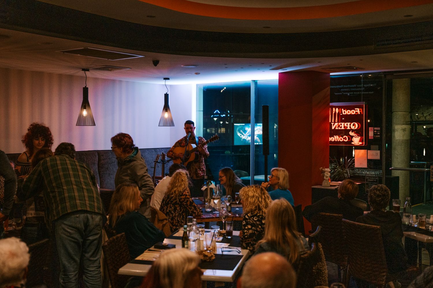 A musician plays an acoustic guitar and sings for patrons seated at tables inside a dimly lit restaurant or cafe at night, with a neon "Open" sign visible in the window