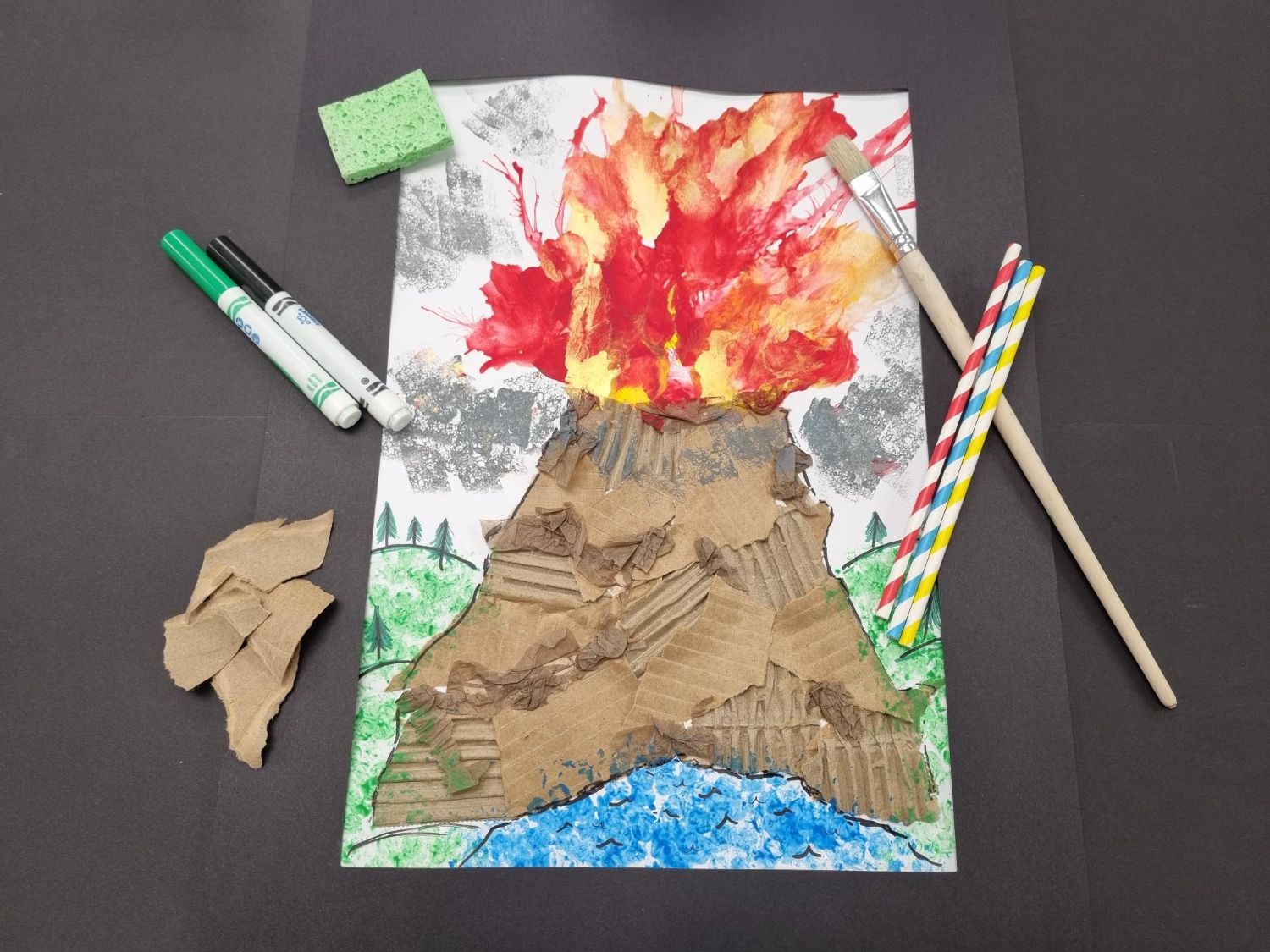 A mixed-media volcano artwork made from torn cardboard and painted red and yellow lava bursts, displayed on a black background with markers, straws, and paintbrushes.