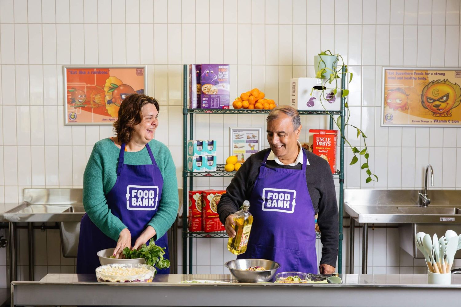 Two smiling people wearing purple Foodbank aprons prepare fresh food at a kitchen counter. A man pours olive oil into a bowl while a woman mixes salad greens.