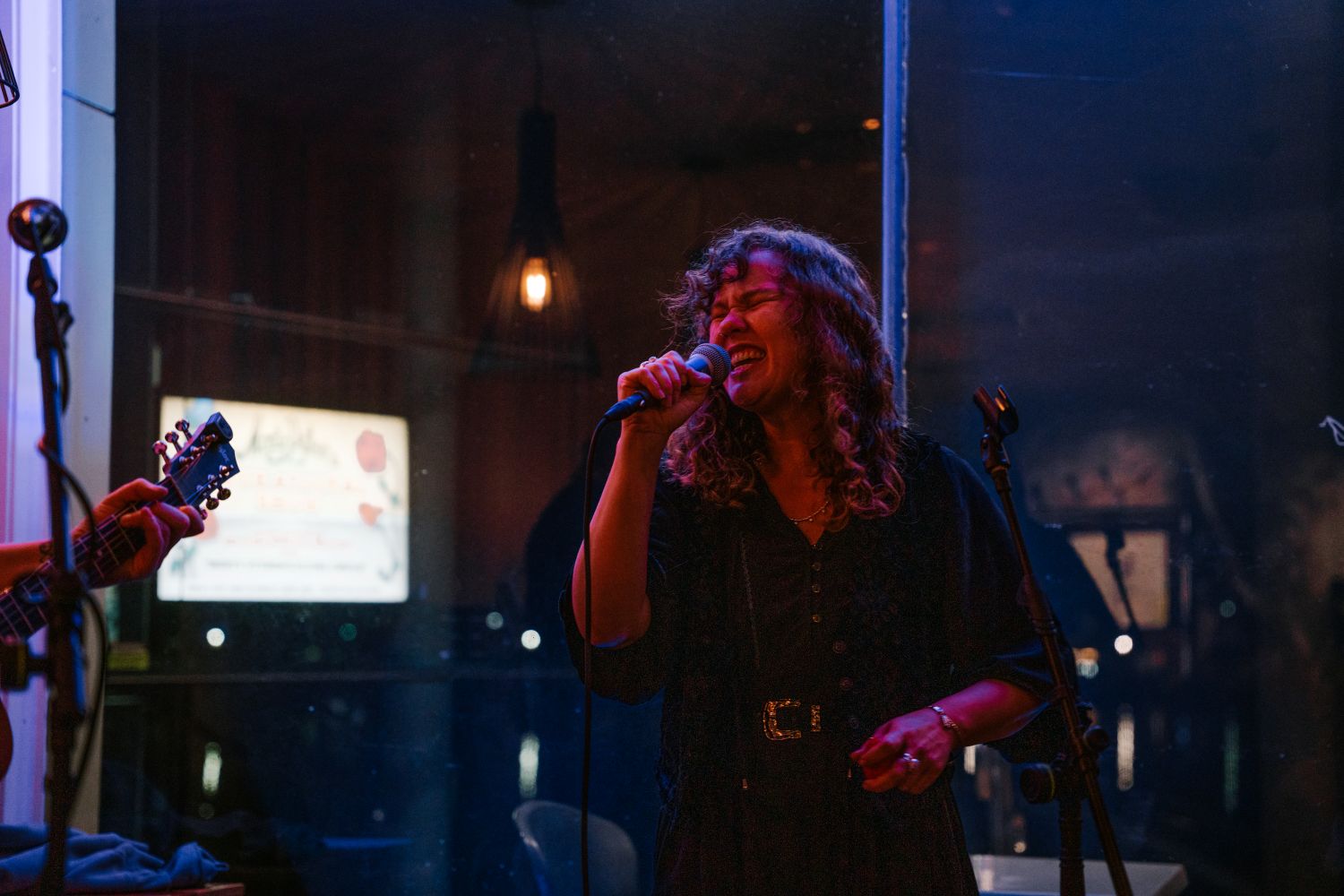 A close-up shot of a female vocalist with curly hair singing passionately into a microphone during a live performance, lit by dramatic blue and red stage lighting.