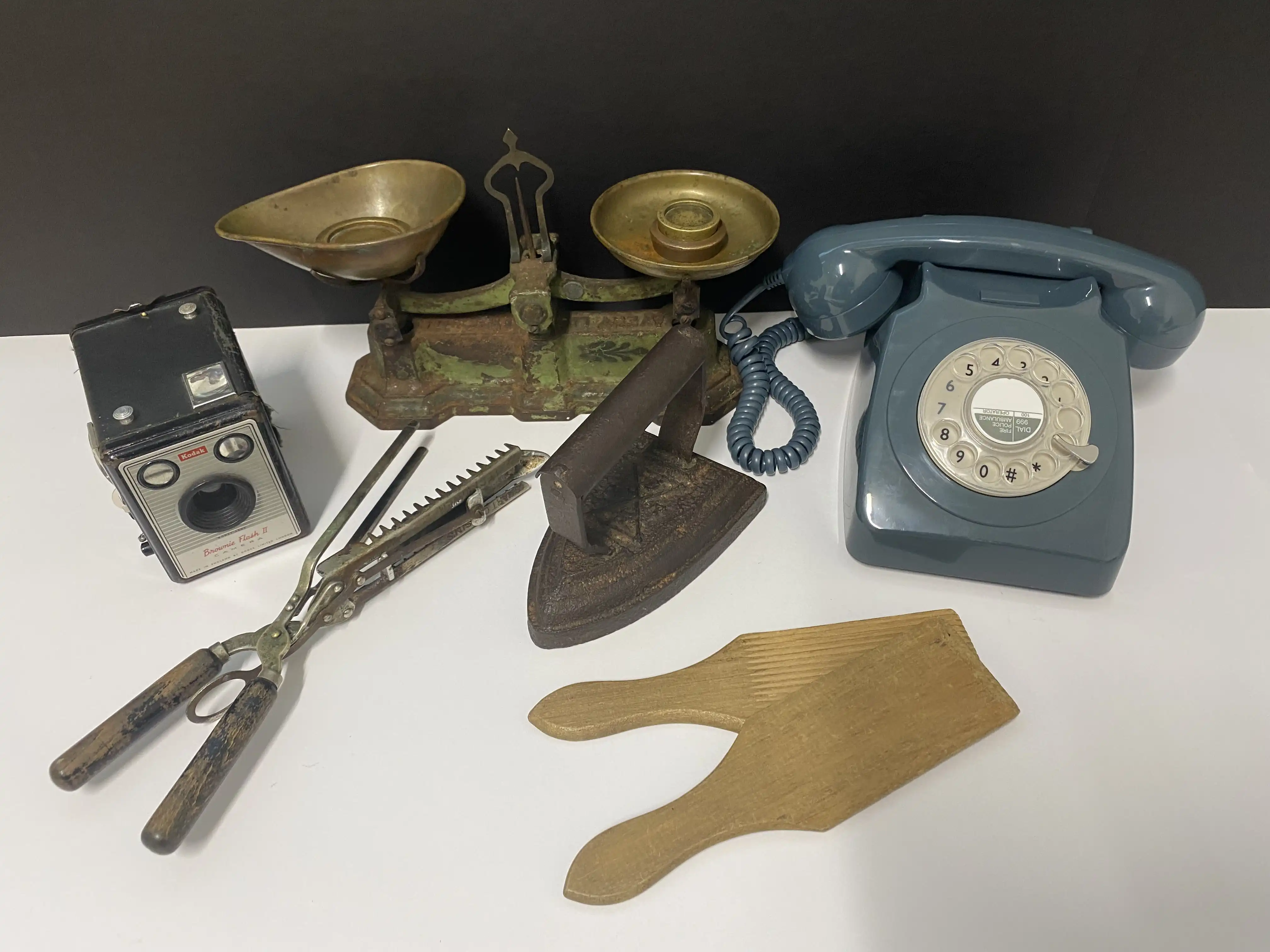 Vintage household items including a rotary phone, balance scale, box camera, scissors, flat iron, and wooden paddles arranged on a table.