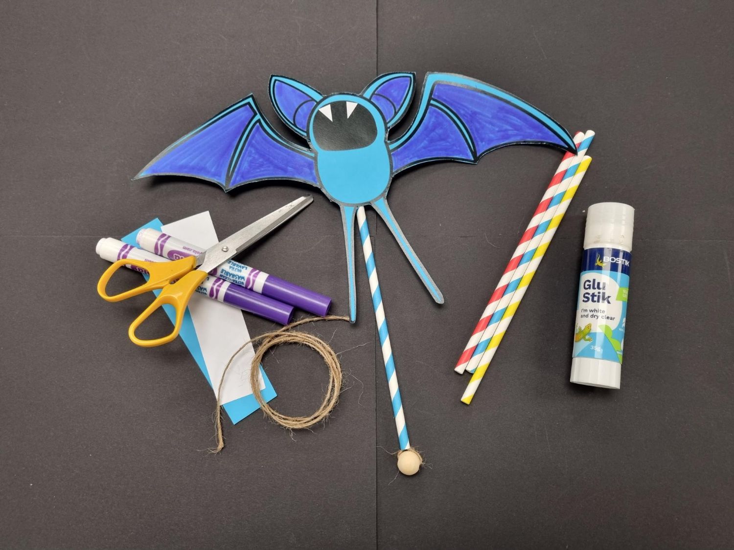 A blue paper Zubat puppet mounted on a straw rests on a black background alongside craft scissors, purple markers, twine, striped straws, and a glue stick.