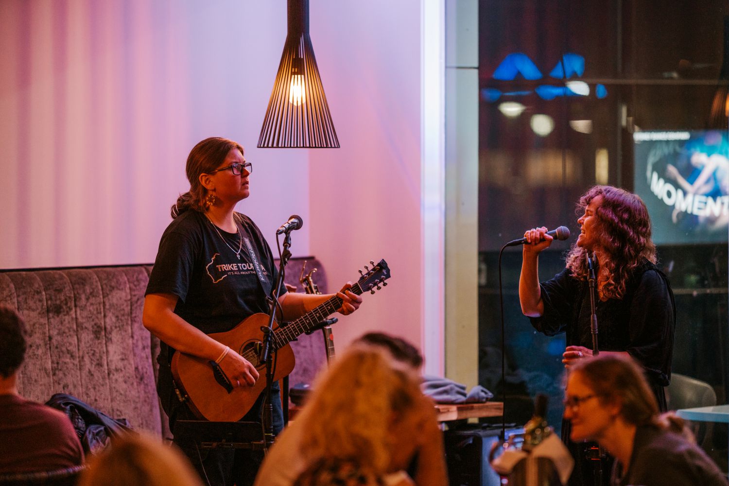 Two women perform live music in a restaurant or venue. One plays an acoustic guitar and sings into a microphone, while the other sings directly into a handheld microphone beside her.