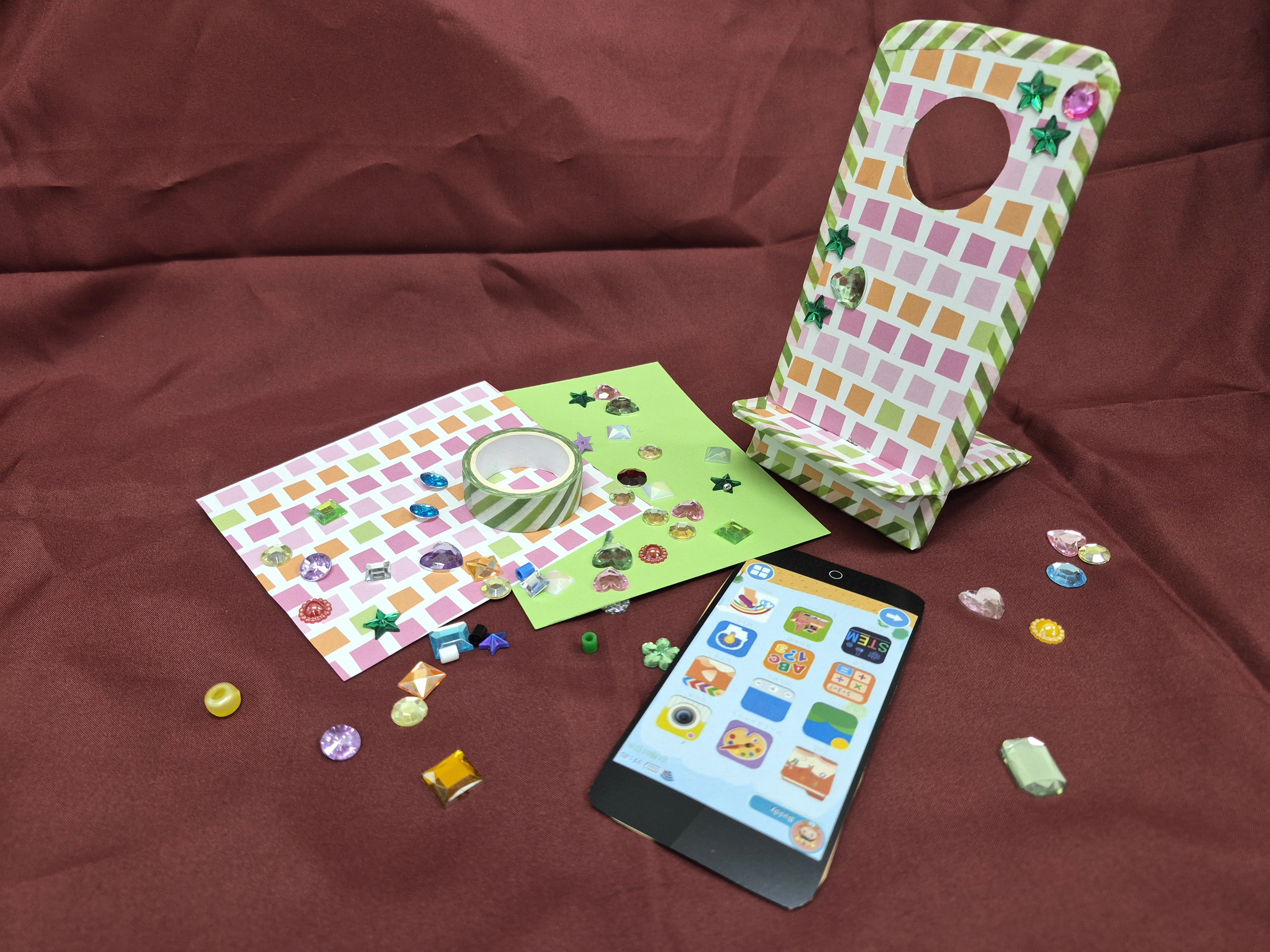 A handmade colourful mobile phone stand sitting on a red velvet background, with a phone lying in front of it and craft stickers.