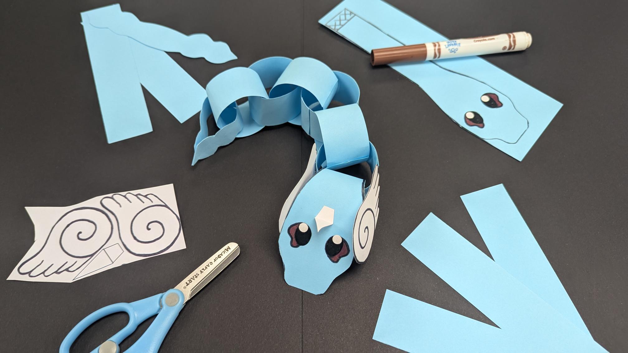 A craft version of a Pokemon Dragonair lying on a black table with scissors and pieces of blue craft paper around it.