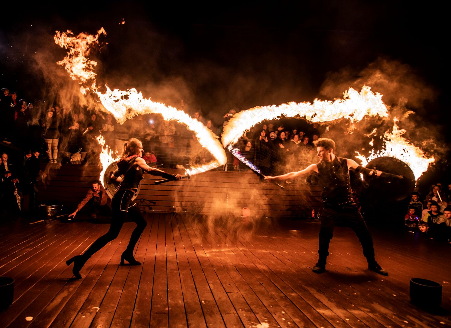 Two fire performers clad in black stage an energetic mock battle on an outdoor wooden deck at night, wielding flaming weapons that leave long, dramatic trails of fire in the air.
