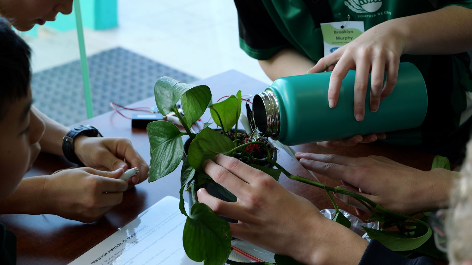 A close-up shot of students. One student pours water from a teal flask into the soil while others adjust wiring connected to a micro:bit sensor