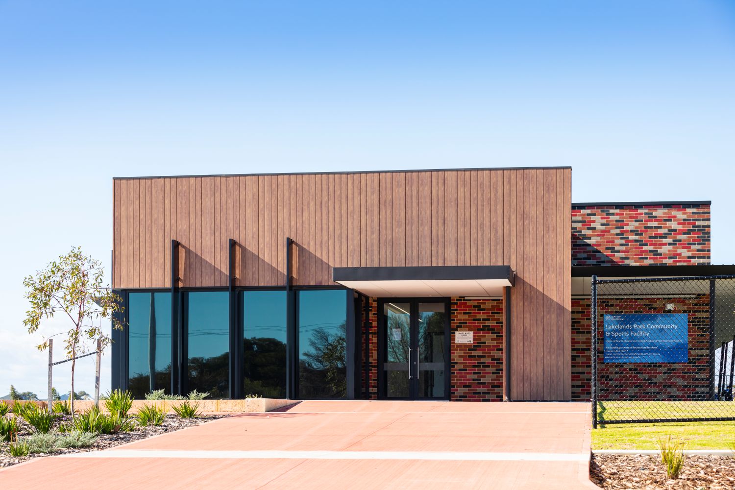 Lakelands Sports Facility exterior showing a modern building with timber-look cladding, brick detailing, large glass windows and a paved entry area.