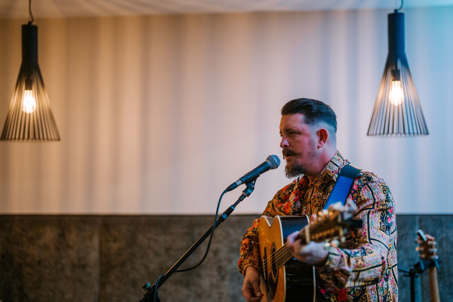 A close-up shot of a male musician with a goatee and mustache singing into a microphone while playing an acoustic guitar, framed by two modern pendant lights in a dimly lit venue.