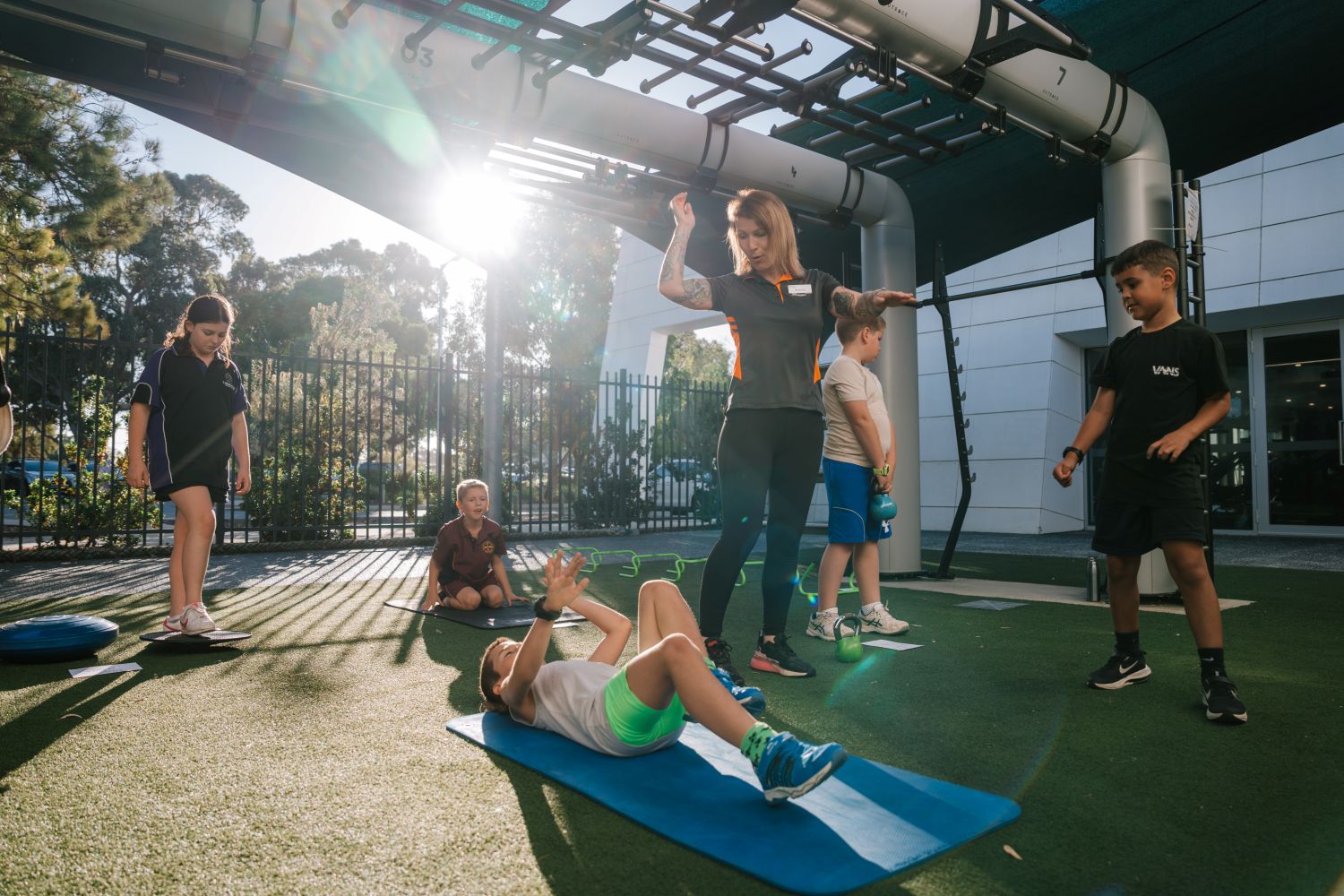 Under a shaded outdoor structure, a female instructor guides a group of children through a fitness circuit including sit-ups on a mat, balancing, and kettlebell lifting on a sunny day.