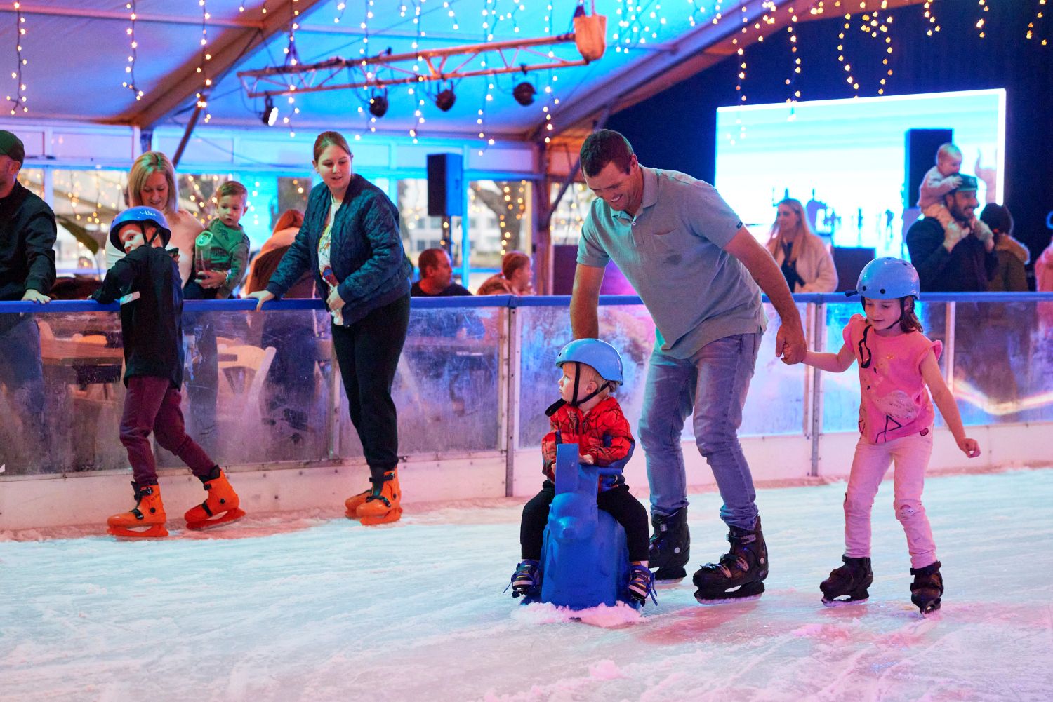 A father skates on an indoor ice rink, guiding his young son who is sitting on a blue plastic skating aid, while his young daughter skates alongside them holding his hand.