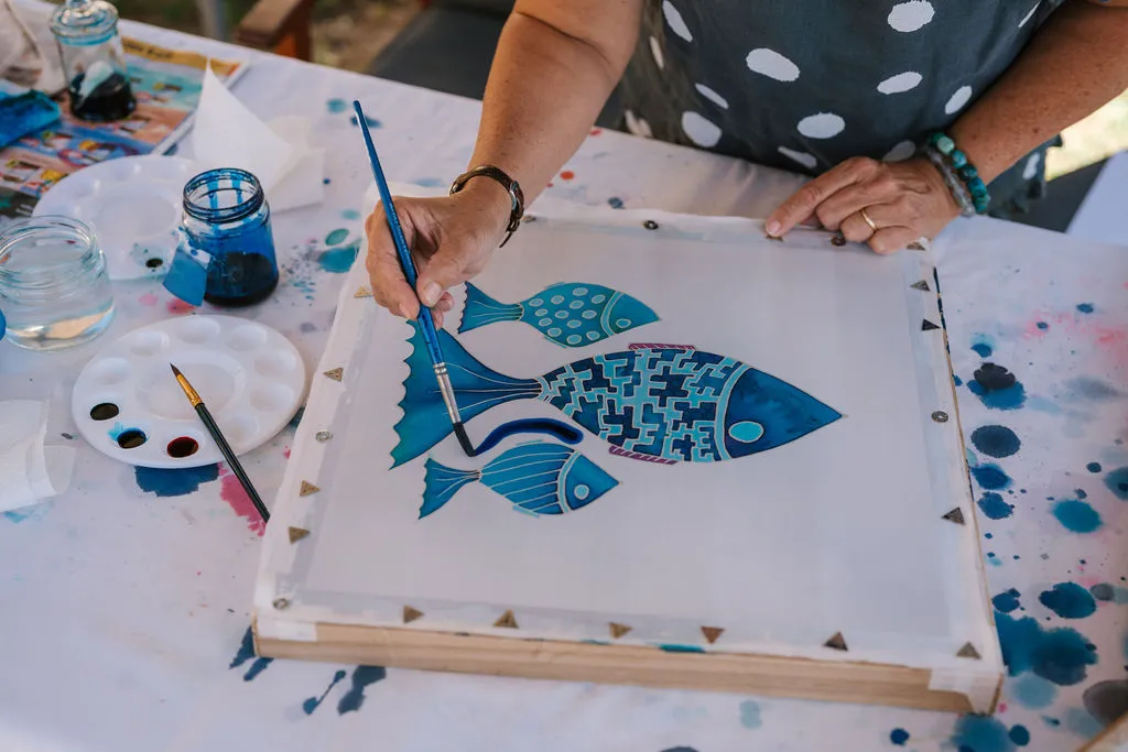 Artist painting on canvas with 3 blue fish with paint brushes and palette next to canvas. 