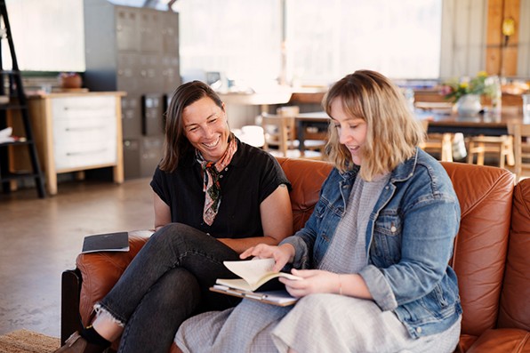 Two women sit next to each other on a couch with happy faces, one looking at the other's notebook